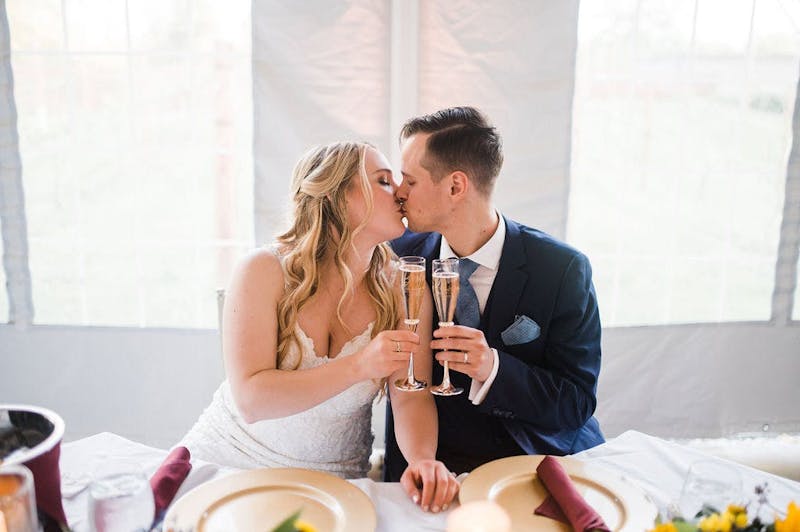 Bride and groom in a deep blue suit, cheersing at the sweetheart table.